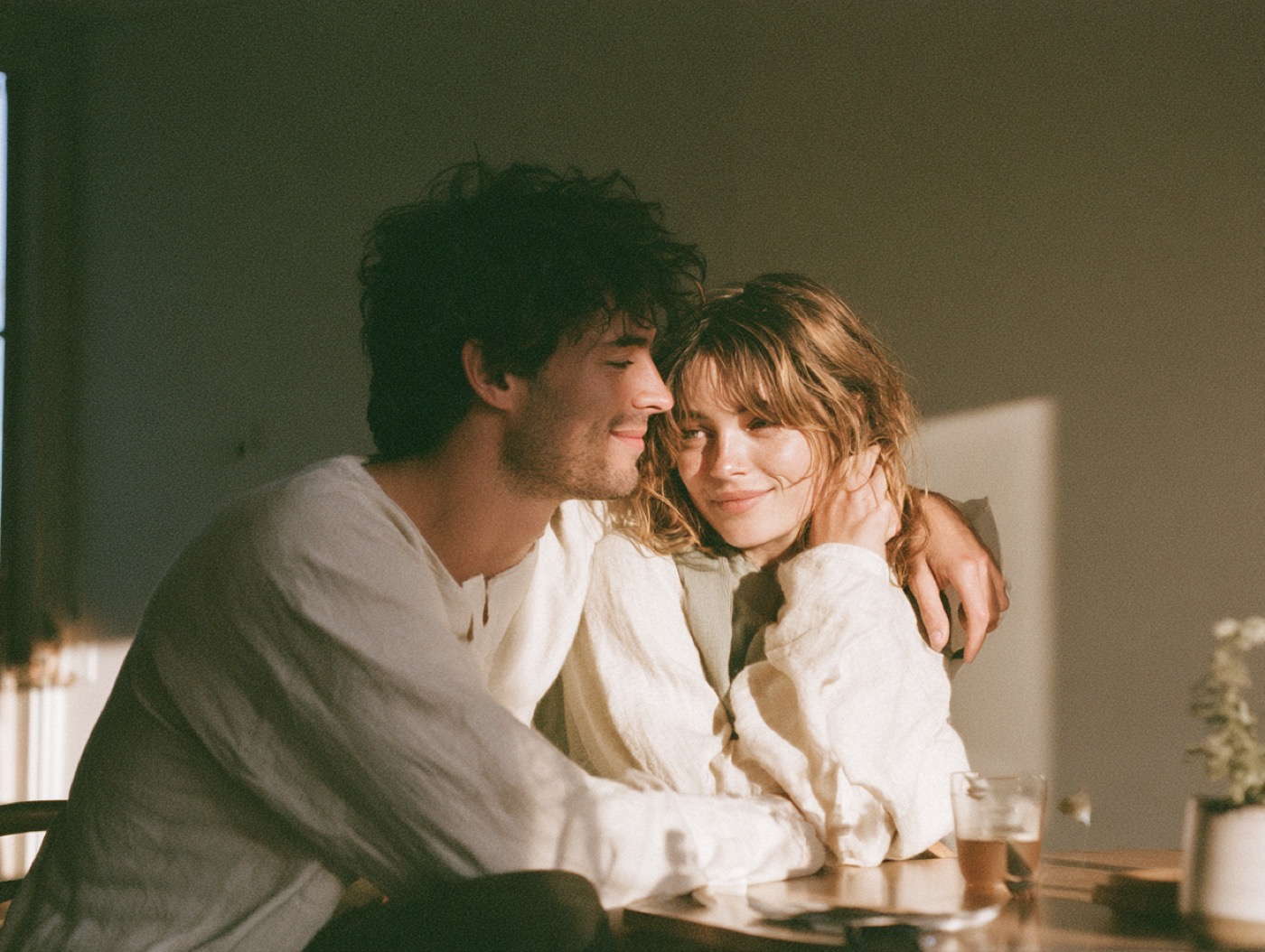 A couple embracing at their kitchen table in warm morning light, close and at ease.
